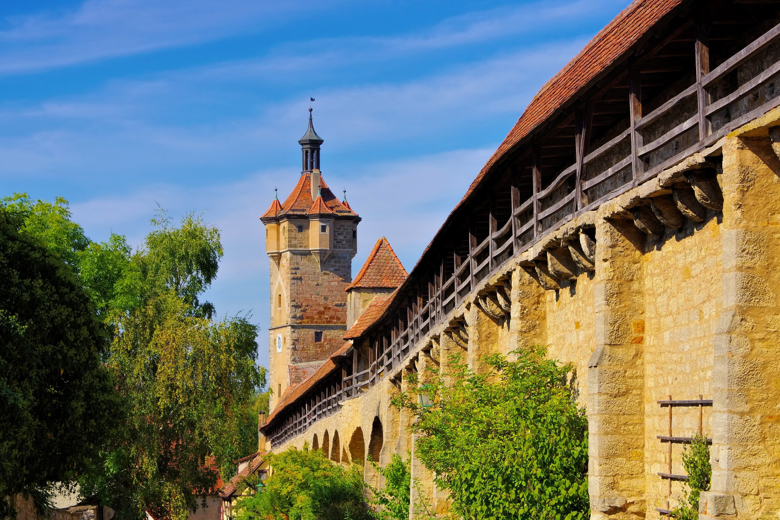 德國羅騰堡(Rothenburg ob der Tauber)的中世紀城牆。圖片顯示一條長長的石牆,上方有有蓋的木製戰鬥走道,背景是一座高聳的塔樓,周圍有綠色的樹木。