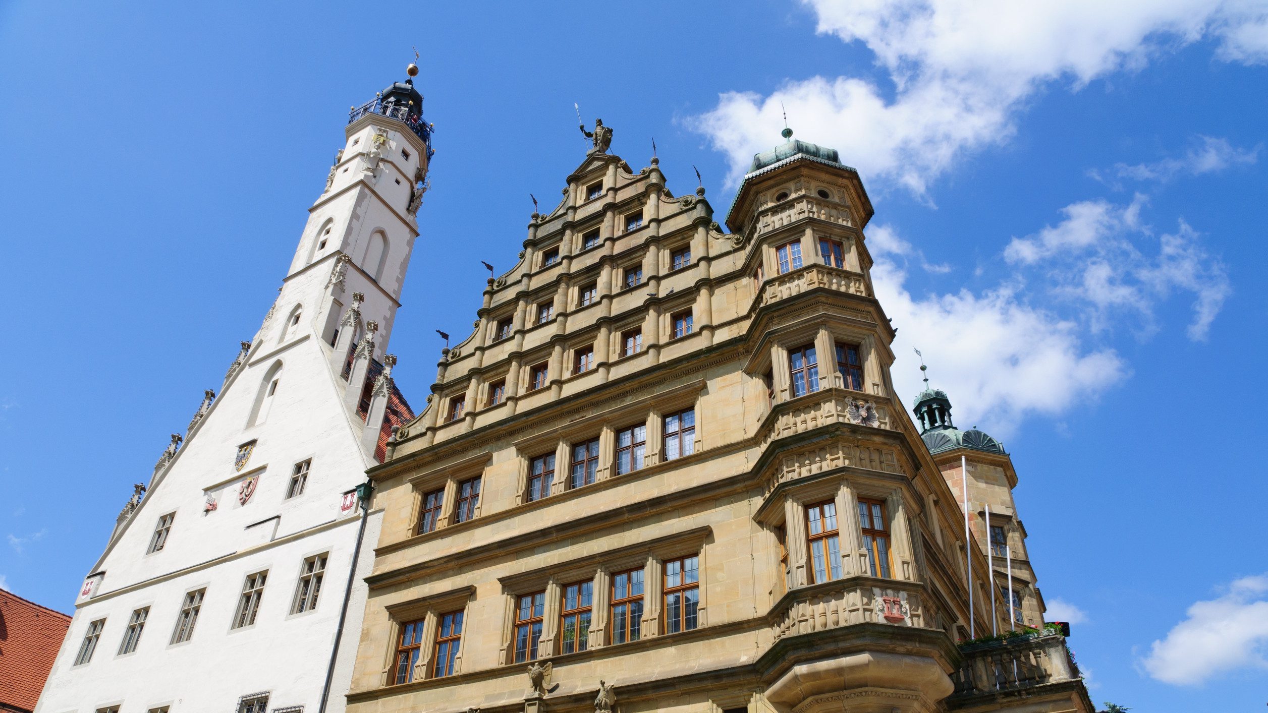 德國羅騰堡(Rothenburg ob der Tauber)的市政廳特寫。畫面左側是高聳的白色哥德式塔樓,右側是米黃色石材、有著多層山形牆的文藝復興風格主體建築,背景是藍天白雲。