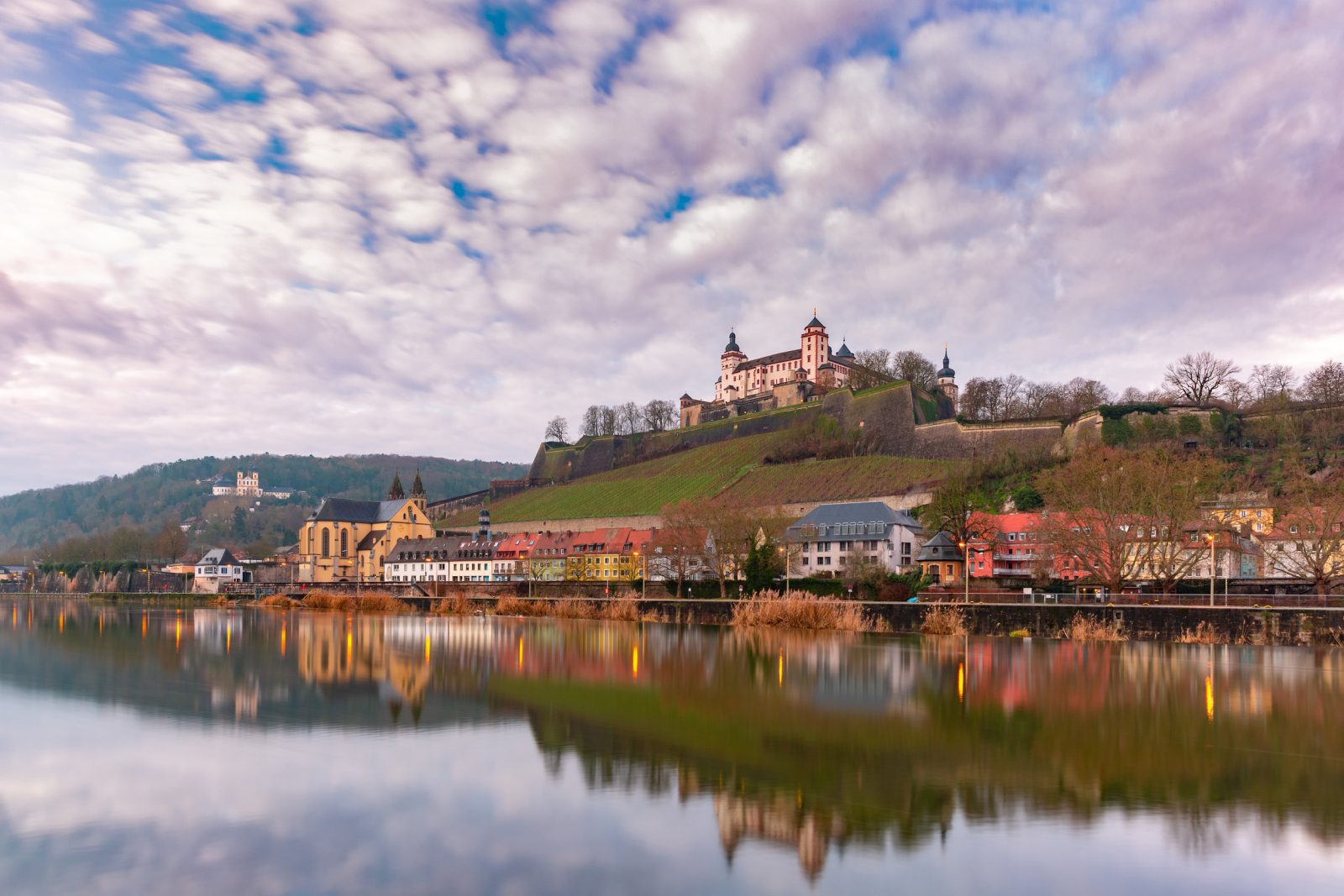 德國維爾茨堡(Würzburg)的景觀。山丘上坐落著雄偉的美因河城堡 (Marienberg Fortress),山腳下是一排沿河的彩色房屋,倒影映在平靜的美因河水面上,天空佈滿雲朵。